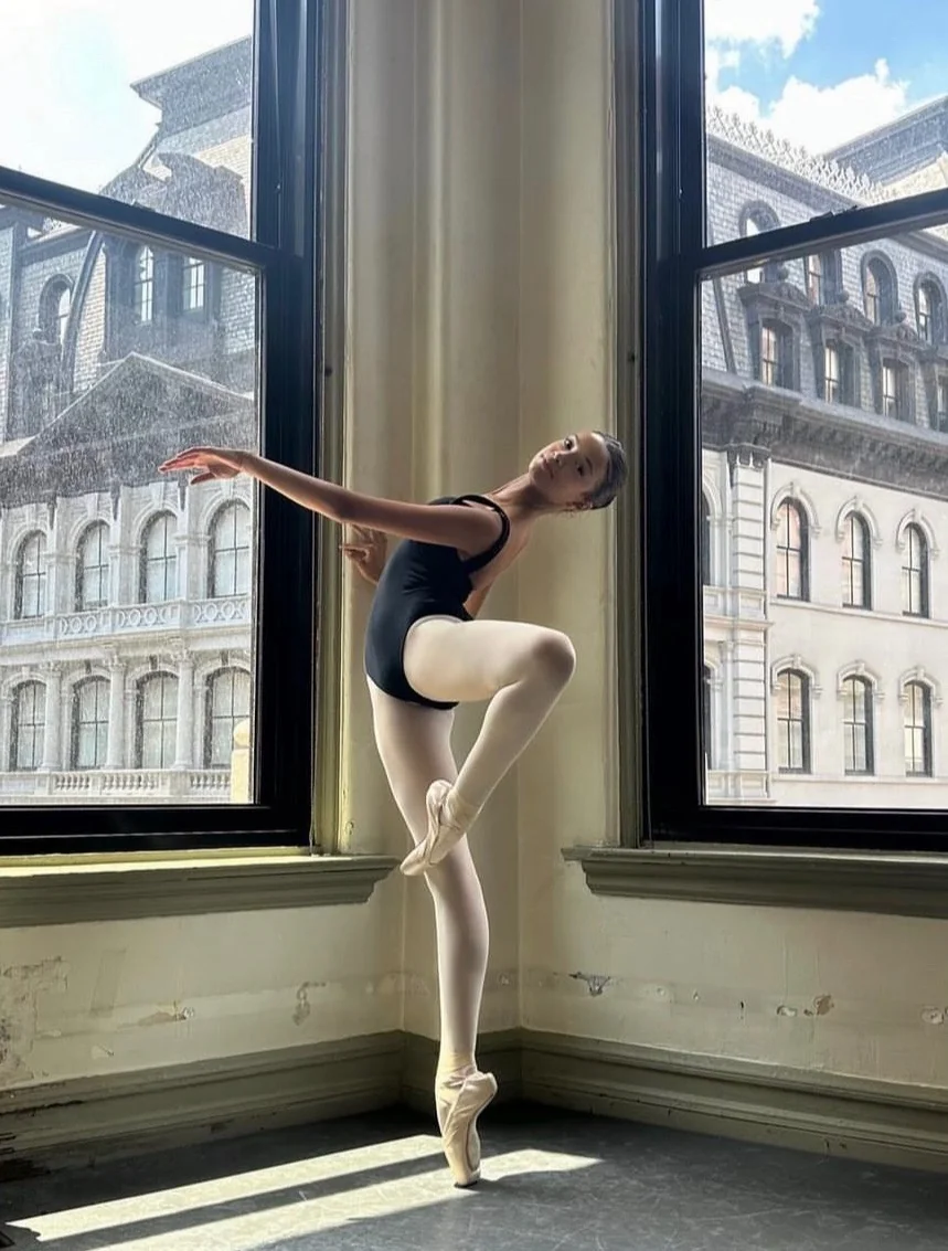 Grace poised en pointe in a black leotard beside sunlit studio windows